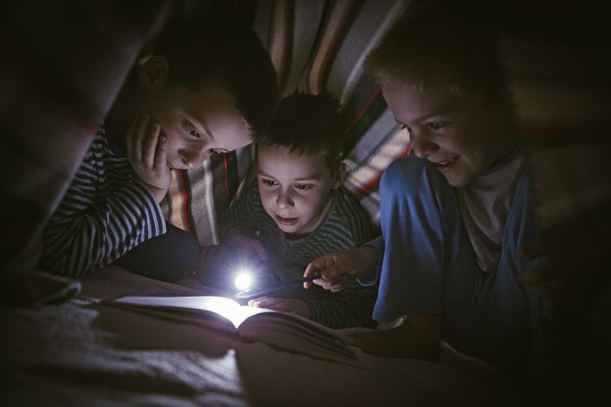 three boys reading under blanket with flashlight